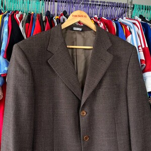 May include: A brown blazer with a subtle checkered pattern, hanging on a wooden hanger. The blazer has two buttons and a notched lapel. The hanger has the text "TOUR SKATE CO." printed on it.