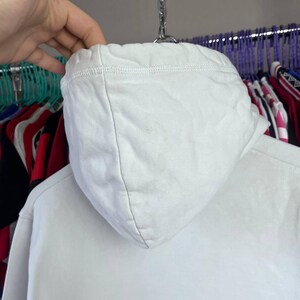 May include: Close-up of a white hoodie hood. The hood is made of a soft, textured fabric and is hanging on a metal hanger. The hood is a solid white colour and appears to be part of a sweatshirt or jacket.