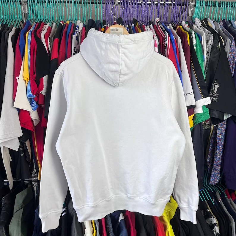 May include: A plain white hoodie with a drawstring hood and long sleeves. The sweatshirt is displayed on a clothes rack, with other garments visible in the background.