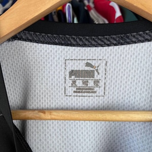 Puede incluir: Una camiseta deportiva blanca con cuello negro y textura de malla. La etiqueta interior del cuello muestra el logotipo de Puma y el texto que indica la talla XXL para EE. UU., TG para Canad&aacute; y 2XL para Europa, impreso en color gris oscuro.