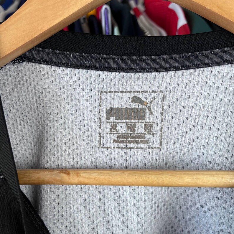 Puede incluir: Una camiseta deportiva blanca con cuello negro y textura de malla. La etiqueta interior del cuello muestra el logotipo de Puma y el texto que indica la talla XXL para EE. UU., TG para Canad&aacute; y 2XL para Europa, impreso en color gris oscuro.