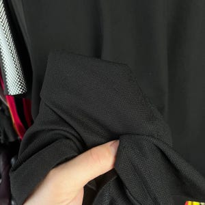 May include: Close-up of a black fabric, possibly a garment. The material appears to be a textured weave, with a visible pattern. The fabric is being held, showing the texture and drape of the material. The background is out of focus.