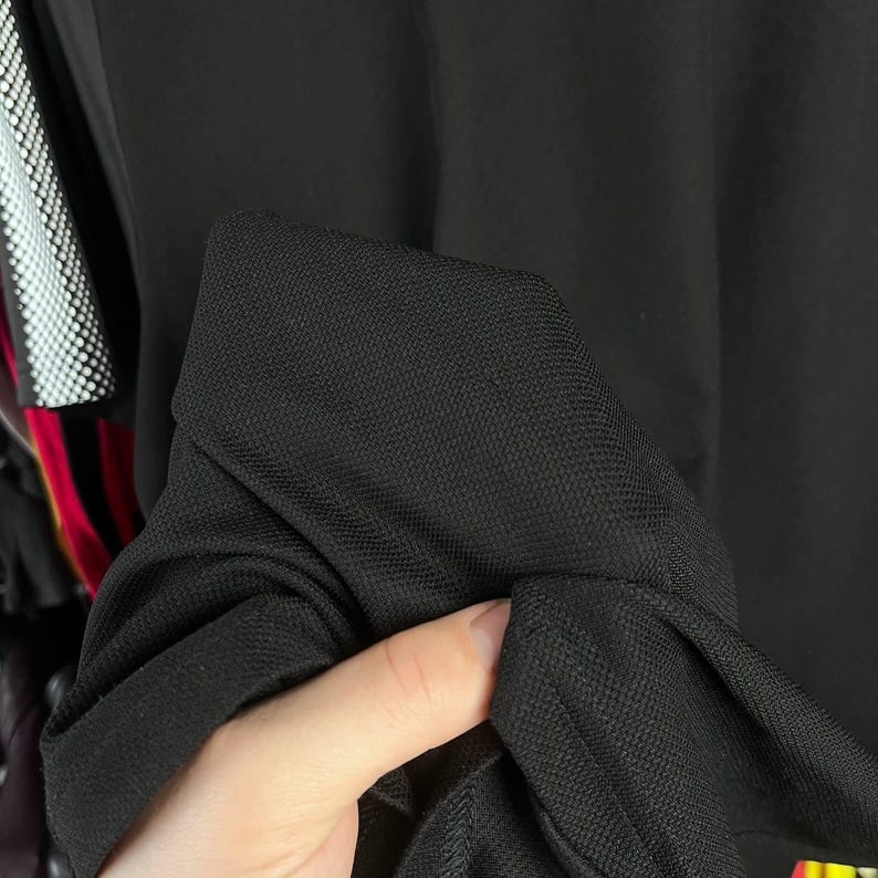 May include: Close-up of a black fabric, possibly a garment. The material appears to be a textured weave, with a visible pattern. The fabric is being held, showing the texture and drape of the material. The background is out of focus.