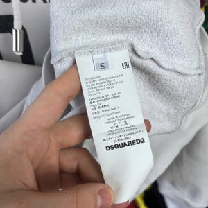 May include: A white garment tag with black text, featuring the brand name DSQUARED2 and size "S". The tag provides details about the product's origin and care. The tag is held by a hand, with the garment's fabric visible.
