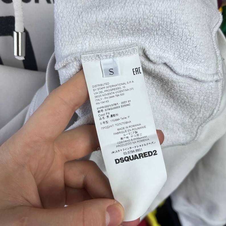 May include: A white garment tag with black text, featuring the brand name DSQUARED2 and size "S". The tag provides details about the product's origin and care. The tag is held by a hand, with the garment's fabric visible.