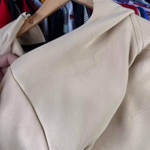 Puede incluir: Primer plano de una chaqueta beige, que muestra el &aacute;rea del hombro y el cuello. La tela parece ser un material liso y tejido. La chaqueta est&aacute; colgada en una percha de madera, con un vistazo de otras prendas en el fondo.