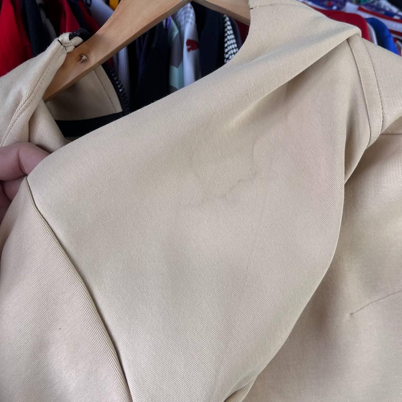 Puede incluir: Primer plano de una chaqueta beige, que muestra el &aacute;rea del hombro y el cuello. La tela parece ser un material liso y tejido. La chaqueta est&aacute; colgada en una percha de madera, con un vistazo de otras prendas en el fondo.
