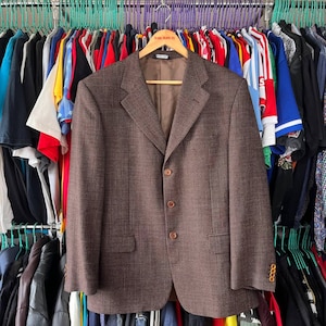 May include: A brown blazer with a subtle checkered pattern, three buttons, and a notched lapel hangs on a wooden hanger. The jacket has two front pockets and buttoned cuffs. The background shows a rack of various clothing items.