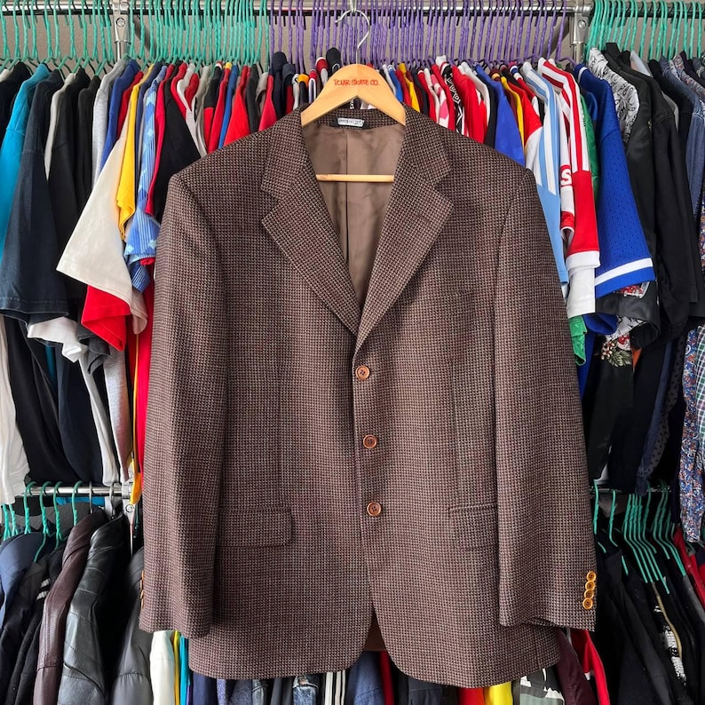 May include: A brown blazer with a subtle checkered pattern, three buttons, and a notched lapel hangs on a wooden hanger. The jacket has two front pockets and buttoned cuffs. The background shows a rack of various clothing items.