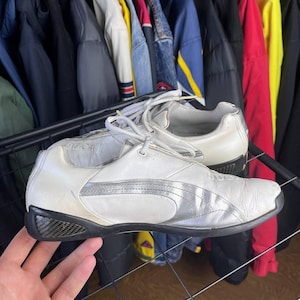 May include: Pair of white Puma sneakers with silver accents and black soles. The shoes have a low-profile design with laces and a textured pattern on the sides. The background shows a rack of clothing.