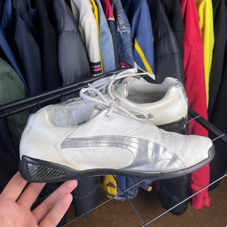 May include: Pair of white Puma sneakers with silver accents and black soles. The shoes have a low-profile design with laces and a textured pattern on the sides. The background shows a rack of clothing.