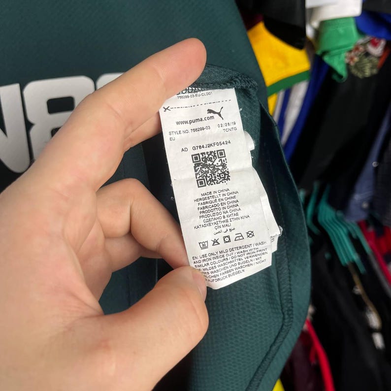 K&ouml;nnte beinhalten: Ein dunkelgr&uuml;nes Sportkleidungsst&uuml;ck mit einem wei&szlig;en Pflegeetikett auf der Innenseite, das das Puma-Logo, die Modellnummer 756299-03, einen QR-Code und Herstellungsdetails zeigt. Der Stoff hat eine texturierte, atmungsaktive Oberfl&auml;che.