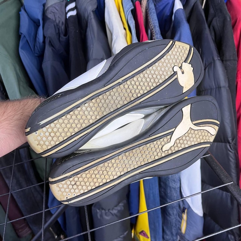 May include: Pair of Puma sneakers with white and gray uppers and black soles. The soles have a tan and black tread pattern with the Puma logo. The sneakers are displayed against a backdrop of hanging clothing.