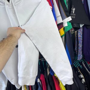 May include: A white hoodie with a drawstring hood and long sleeves. The sleeve's texture is visible. The background shows various garments hanging on rails. The hoodie is held by a hand.