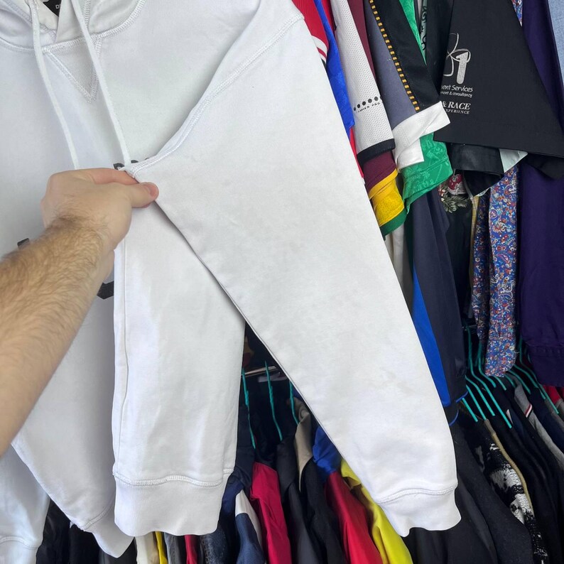 May include: A white hoodie with a drawstring hood and long sleeves. The sleeve's texture is visible. The background shows various garments hanging on rails. The hoodie is held by a hand.