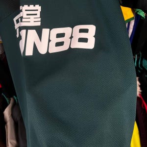 K&ouml;nnte beinhalten: Ein dunkelgr&uuml;nes Sporttrikot mit wei&szlig;em Aufdruck, der FUN88 sowie chinesische Schriftzeichen zeigt. Der Stoff weist eine dezent texturierte Oberfl&auml;che auf und ist zusammen mit anderen Kleidungsst&uuml;cken in einem Schrank zu sehen.