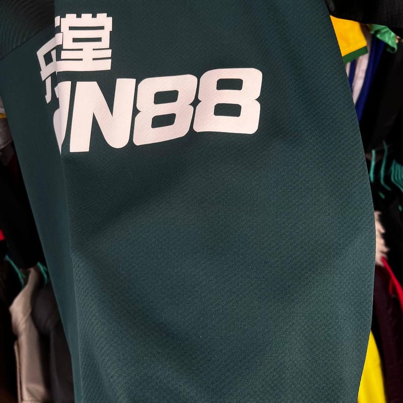 K&ouml;nnte beinhalten: Ein dunkelgr&uuml;nes Sporttrikot mit wei&szlig;em Aufdruck, der FUN88 sowie chinesische Schriftzeichen zeigt. Der Stoff weist eine dezent texturierte Oberfl&auml;che auf und ist zusammen mit anderen Kleidungsst&uuml;cken in einem Schrank zu sehen.