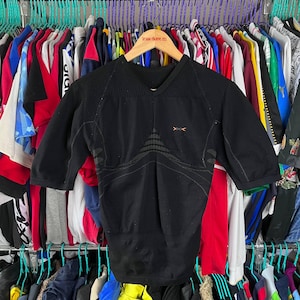 Op de afbeelding: Zwart sportshirt met korte mouwen en een V-hals. Het shirt heeft een textuurontwerp op de schouders en een geometrisch patroon op de borst. Kleine oranje pijlachtige symbolen zijn zichtbaar bij de halslijn. Het shirt hangt aan een houten hanger.