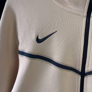 Puede incluir: Chaqueta con cremallera beige claro con el logotipo de Nike en azul oscuro y un dise&ntilde;o de rayas a juego en el pecho. La chaqueta tiene una cremallera completa y est&aacute; hecha de una tela suave y texturizada.