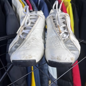 May include: Pair of white and silver Ducati sneakers with black soles. The word "DUCATI" is embossed on the side of each shoe. The sneakers have white laces and show signs of wear.