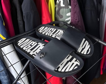 May include: Black slide sandals with a white and black patterned footbed. The sandals have a wide strap and a small silver-colored emblem. The sandals are displayed on a black metal rack, with clothing in the background.