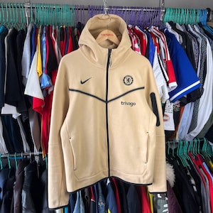 Puede incluir: Sudadera con capucha y cremallera de color beige con detalles negros y el logo del Chelsea. Presenta el logo de Nike y la palabra "trivago" en negro. La sudadera est&aacute; colgada en una percha con otras prendas.