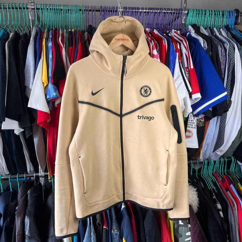 Puede incluir: Sudadera con capucha y cremallera de color beige con detalles negros y el logo del Chelsea. Presenta el logo de Nike y la palabra "trivago" en negro. La sudadera est&aacute; colgada en una percha con otras prendas.