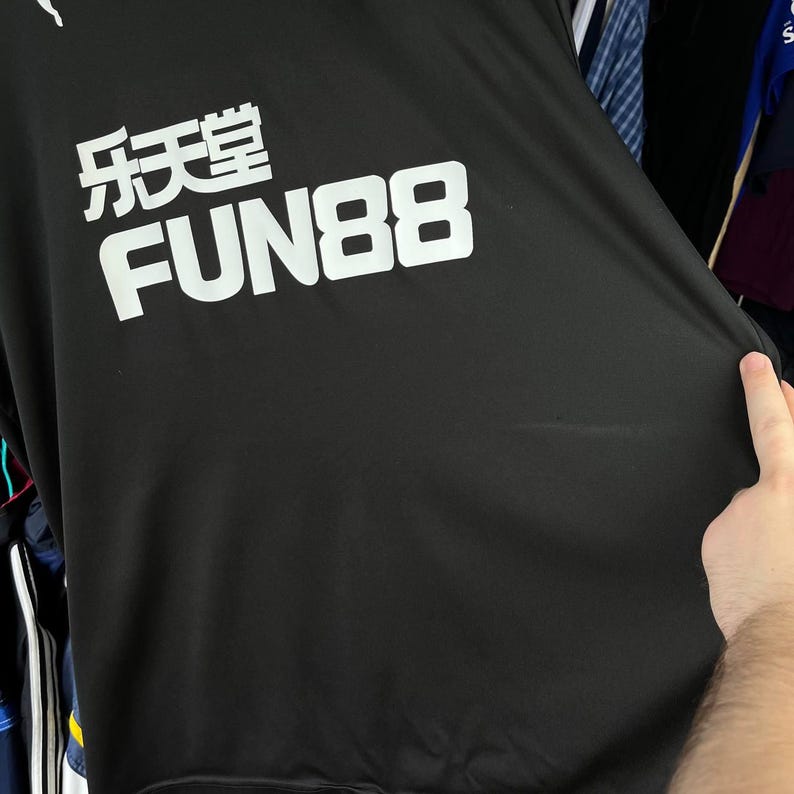 Puede incluir: Una camiseta deportiva negra con el texto blanco FUN88 y caracteres chinos impresos en la parte delantera. La tela es lisa y oscura, con un logotipo blanco visible en el borde superior. La prenda se sostiene para mostrar el dise&ntilde;o gr&aacute;fico.