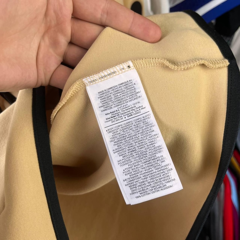 Puede incluir: Primer plano de una camiseta deportiva beige con ribete negro alrededor del cuello y las mangas. Una etiqueta blanca con texto negro est&aacute; cosida en el cuello de la camiseta. La etiqueta incluye informaci&oacute;n sobre los materiales y el origen de la camiseta.