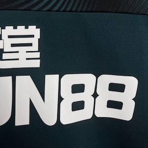 K&ouml;nnte beinhalten: Ein dunkelblaugr&uuml;nes Kleidungsst&uuml;ck aus Stoff mit einem auff&auml;lligen wei&szlig;en Grafikdruck. Das Design enth&auml;lt den Text "FUN88" in gro&szlig;en, blockartigen, serifenlosen Gro&szlig;buchstaben unter einem teilweisen wei&szlig;en chinesischen Schriftzeichen.