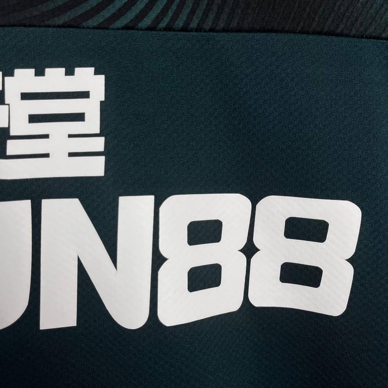 K&ouml;nnte beinhalten: Ein dunkelblaugr&uuml;nes Kleidungsst&uuml;ck aus Stoff mit einem auff&auml;lligen wei&szlig;en Grafikdruck. Das Design enth&auml;lt den Text "FUN88" in gro&szlig;en, blockartigen, serifenlosen Gro&szlig;buchstaben unter einem teilweisen wei&szlig;en chinesischen Schriftzeichen.
