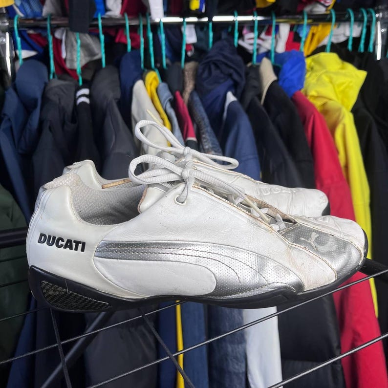 May include: White and silver Puma Ducati sneakers with black soles. The word "DUCATI" is printed on the side of the shoe. The sneakers are on a black metal rack with a background of hanging jackets.