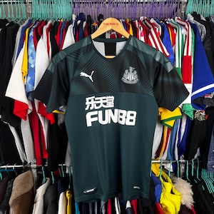 K&ouml;nnte beinhalten: Ein dunkelgr&uuml;nes und schwarzes kurz&auml;rmeliges Fu&szlig;balltrikot mit V-Ausschnitt, dem wei&szlig;en Puma-Logo auf der rechten Brust, dem Newcastle United-Wappen auf der linken Brust und dem wei&szlig;en Schriftzug FUN88 mit chinesischen Schriftzeichen vorne.