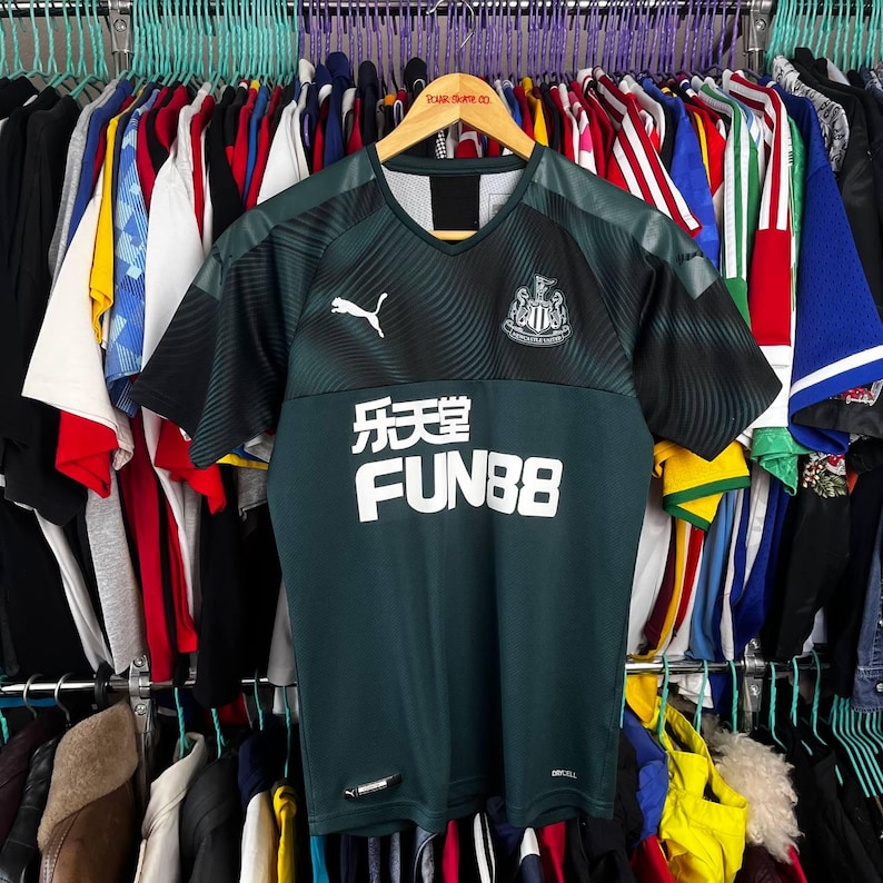 K&ouml;nnte beinhalten: Ein dunkelgr&uuml;nes und schwarzes kurz&auml;rmeliges Fu&szlig;balltrikot mit V-Ausschnitt, dem wei&szlig;en Puma-Logo auf der rechten Brust, dem Newcastle United-Wappen auf der linken Brust und dem wei&szlig;en Schriftzug FUN88 mit chinesischen Schriftzeichen vorne.