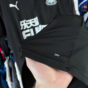 Puede incluir: Una camiseta deportiva negra con un logotipo de Puma blanco, el escudo del Newcastle United y el texto blanco FUN88. La tela incluye una peque&ntilde;a etiqueta plateada con el texto DRYCELL impreso en el dobladillo inferior.
