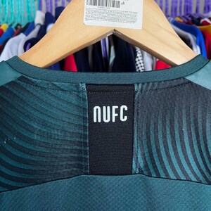K&ouml;nnte beinhalten: Ein dunkelgr&uuml;nes Sporttrikot mit einem schwarzen vertikalen Einsatz in der Mitte des Halsausschnitts, auf dem der wei&szlig;e Text NUFC aufgedruckt ist. Der Stoff zeigt ein dezentes, wellenf&ouml;rmiges, dunkles Muster im Schulter- und Brustbereich.