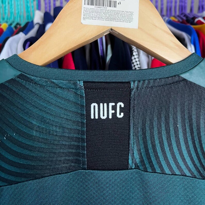 K&ouml;nnte beinhalten: Ein dunkelgr&uuml;nes Sporttrikot mit einem schwarzen vertikalen Einsatz in der Mitte des Halsausschnitts, auf dem der wei&szlig;e Text NUFC aufgedruckt ist. Der Stoff zeigt ein dezentes, wellenf&ouml;rmiges, dunkles Muster im Schulter- und Brustbereich.