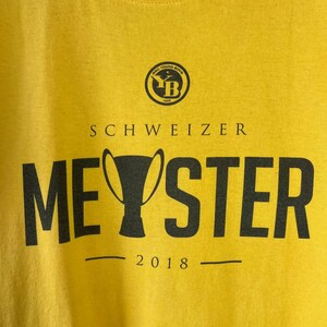 Puede incluir: Camiseta amarilla con el texto negro "SCHWEIZER MEYSTER 2018". Encima, un logotipo circular con las letras "YB" y las palabras "BSC YOUNG BOYS 1898".