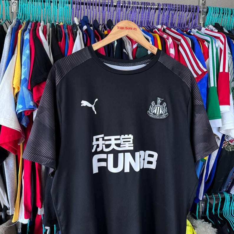 Puede incluir: Una camiseta deportiva negra de manga corta con un logotipo blanco de Puma en el pecho derecho y el escudo del Newcastle United en el izquierdo. El centro muestra el texto blanco FUN88 y caracteres chinos. Las mangas tienen un patr&oacute;n gris texturizado.