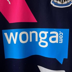 Pu&ograve; includere: Primo piano di una maglia sportiva blu navy con un design rosa e bianco. La maglia presenta la scritta "wonga.com" in bianco su un fumetto blu. Lo stemma del Newcastle United &egrave; visibile.