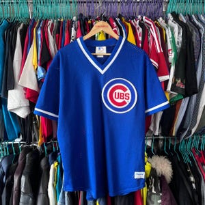 Puede incluir: Camiseta de béisbol azul de los Cachorros de Chicago con ribete blanco en el cuello en V y las mangas. La camiseta presenta el logotipo de los Cachorros en el pecho. La camiseta está colgada en una percha con otras prendas.