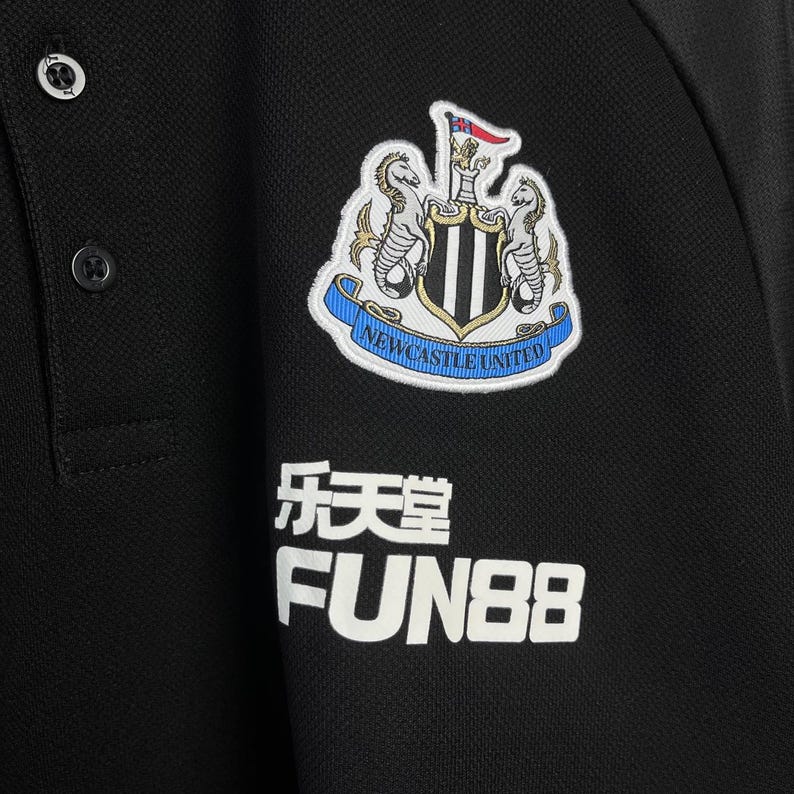 May include: Black polo shirt featuring the Newcastle United crest on the chest. The crest includes two seahorses, a shield, and the words "Newcastle United". Below the crest are Chinese characters and the word "FUN88".