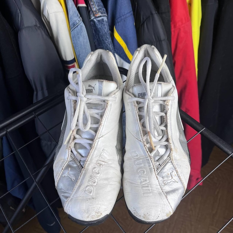 May include: Pair of white Puma Ducati sneakers with silver accents. The shoes have white laces and the word "Ducati" is embossed on the side. The Puma logo is visible on the tongue and side of the shoe. The sneakers are on a metal rack.