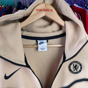 Puede incluir: Sudadera con capucha con cremallera Nike color beige con detalles negros y un parche del Chelsea Football Club. Cuenta con un logotipo de Nike negro y el texto "POLAR SKATE CO." en la percha de madera. La sudadera tiene cremallera completa y capucha.