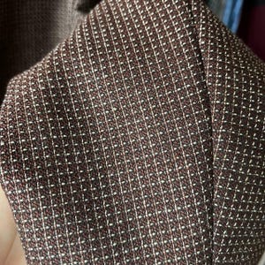 May include: Close-up of a brown fabric with a small white grid pattern. The fabric appears to be part of a garment, possibly a blazer or jacket. The texture is detailed, showing the weave of the material.