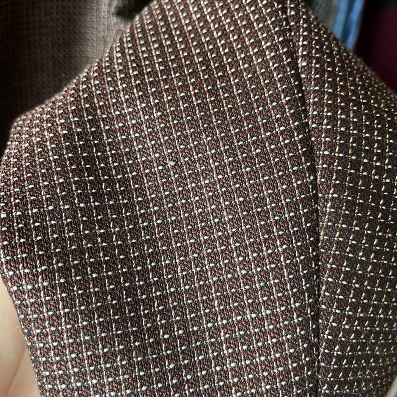 May include: Close-up of a brown fabric with a small white grid pattern. The fabric appears to be part of a garment, possibly a blazer or jacket. The texture is detailed, showing the weave of the material.