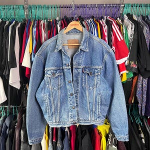 May include: A light blue denim jacket with a classic design, featuring button closures, chest pockets, and a collar. The jacket has a worn look and a brown leather patch on the back. It is hanging on a wooden hanger among other clothing items.