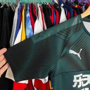 K&ouml;nnte beinhalten: Ein dunkelgr&uuml;nes kurz&auml;rmeliges Sporttrikot mit einem schwarzen Wellenlinienmuster auf den Schultern und &Auml;rmeln. Das Kleidungsst&uuml;ck zeigt ein wei&szlig;es Puma-Logo auf der Brust und einen teilweisen wei&szlig;en Grafikdruck mit chinesischen Schriftzeichen unten.