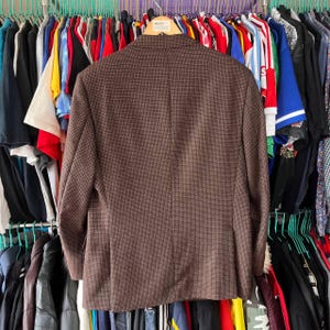 May include: A brown blazer with a subtle checkered pattern, hanging on a rack with other clothing items. The blazer has a classic design, with a notched lapel and a two-button closure. The fabric appears to be a wool blend.