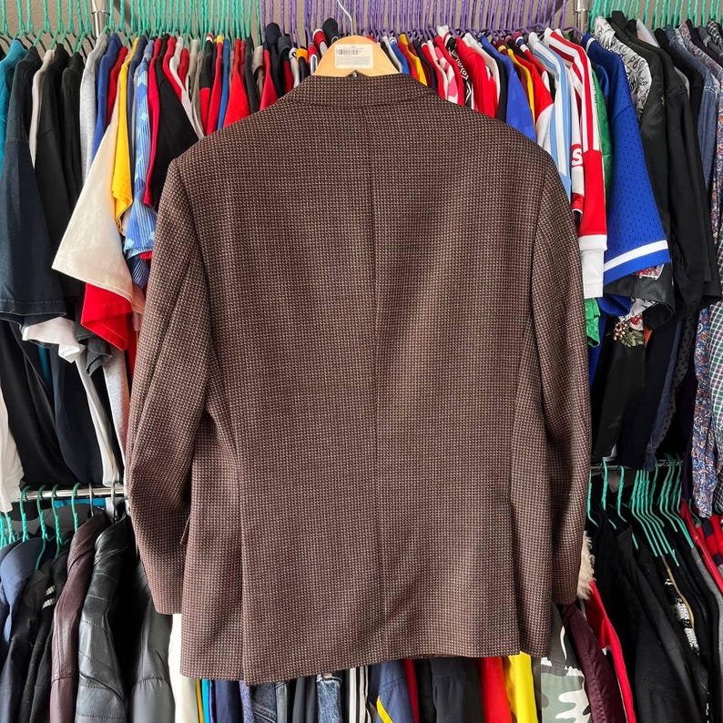 May include: A brown blazer with a subtle checkered pattern, hanging on a rack with other clothing items. The blazer has a classic design, with a notched lapel and a two-button closure. The fabric appears to be a wool blend.